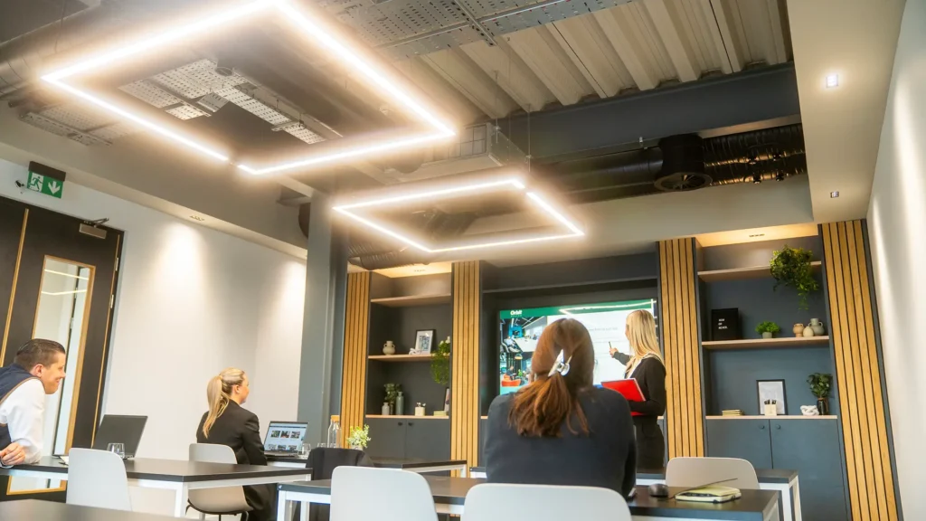 Office space with square LED ceiling lights, people working at desks, and a video call on a screen.