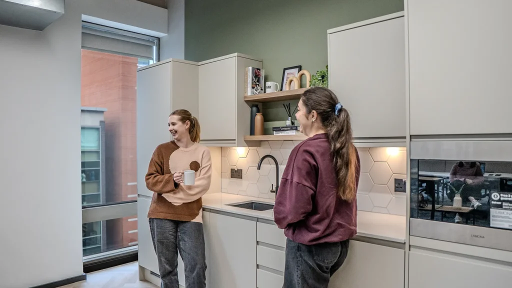 Two women chatting in a small office kitchenette with white cabinetry and a green accent wall.